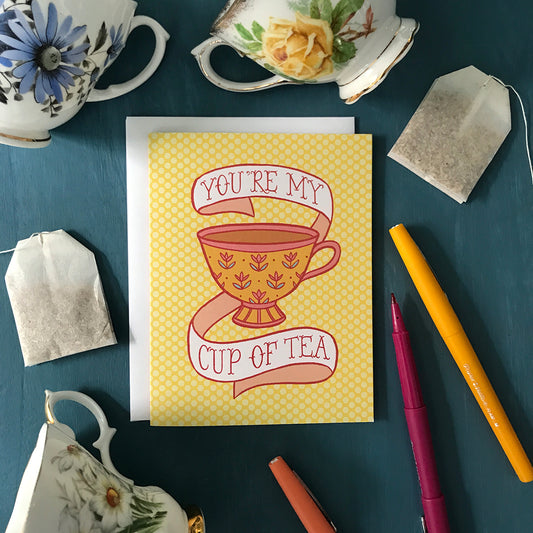 A yellow polka dot greeting card features an illustration of a golden teacup with yellow flowers, full of strong tea. Around the teacup is a banner that reads You're my cup of tea in pink letters. The card is surrounded by floral china teacups, felt-tip pens, and teabags.⁠