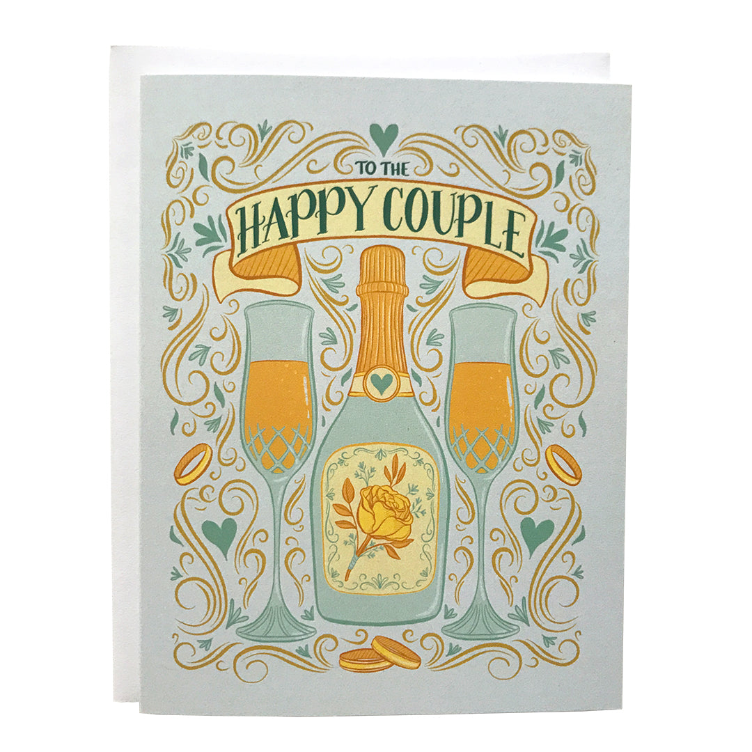 A pastel blue greeting card features an illustration of two full champagne flutes, a champagne bottle with a yellow rose, wedding bands, Art Nouveau flourishes, hearts, and the words To The Happy Couple. The card sits against a white envelope on a white background.