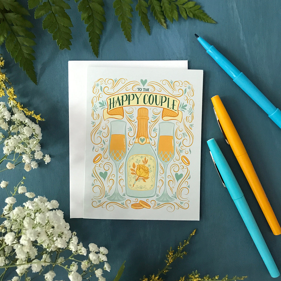 A pastel blue greeting card features an illustration of two full champagne flutes, a champagne bottle with a yellow rose, wedding bands, Art Nouveau flourishes, hearts, and the words To The Happy Couple. The card is flanked by greenery and pens.