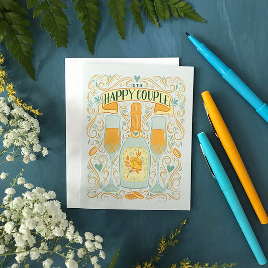 A pastel blue greeting card features an illustration of two full champagne flutes, a champagne bottle with a yellow rose, wedding bands, Art Nouveau flourishes, hearts, and the words To The Happy Couple. The card is flanked by greenery and pens.