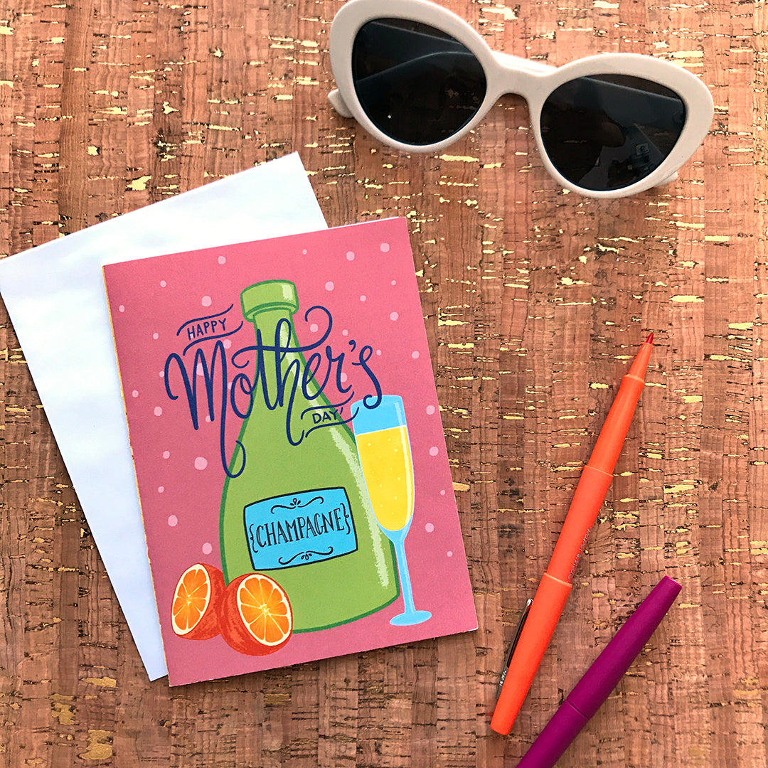 A pink greeting card reads Happy Mother's Day in blue hand lettering on top of an illustration of a green bottle of champagne, an orange cut in half, and a full champagne flute. The card rests on a cork background, and around it are a pair of sunglasses and orange and pink pens.