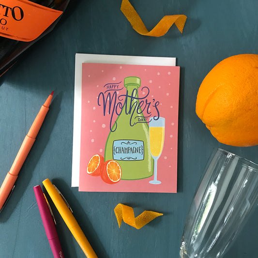 A pink greeting card reads Happy Mother's Day in blue hand lettering on top of an illustration of a green bottle of champagne, an orange cut in half, and a full champagne flute. The card rests on a blue background, and around it are an orange, two orange twists, a champagne flute, orange, pink and yellow pens, and the edge of a bottle of champagne.
