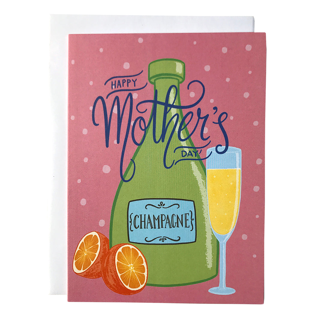 A pink greeting card reads Happy Mother's Day in blue hand lettering on top of an illustration of a green bottle of champagne, an orange cut in half, and a full champagne flute. The card sits against a white envelope on a white background.