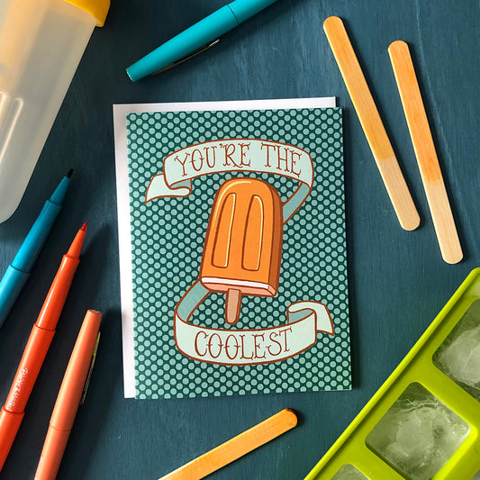 A turquoise polka dot greeting card shows an illustration of a creamy orange popsicle surrounded by a banner that reads you're the coolest in hand lettering. The card sits on a blue wooden background and is surrounded by a filled green ice cube tray, orange, red and blue pens, a plastic popsicle mold and popsicle sticks dyed orange.