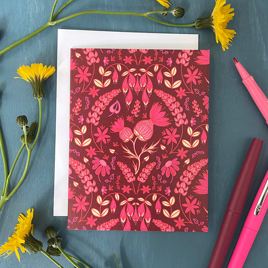 A dark pink greeting card is covered in a folk-style pattern of pink thistles, lupines, haskap berries and mayflowers, with creamy yellow leafy accents. The card is flanked by yellow flowers and pink pens.