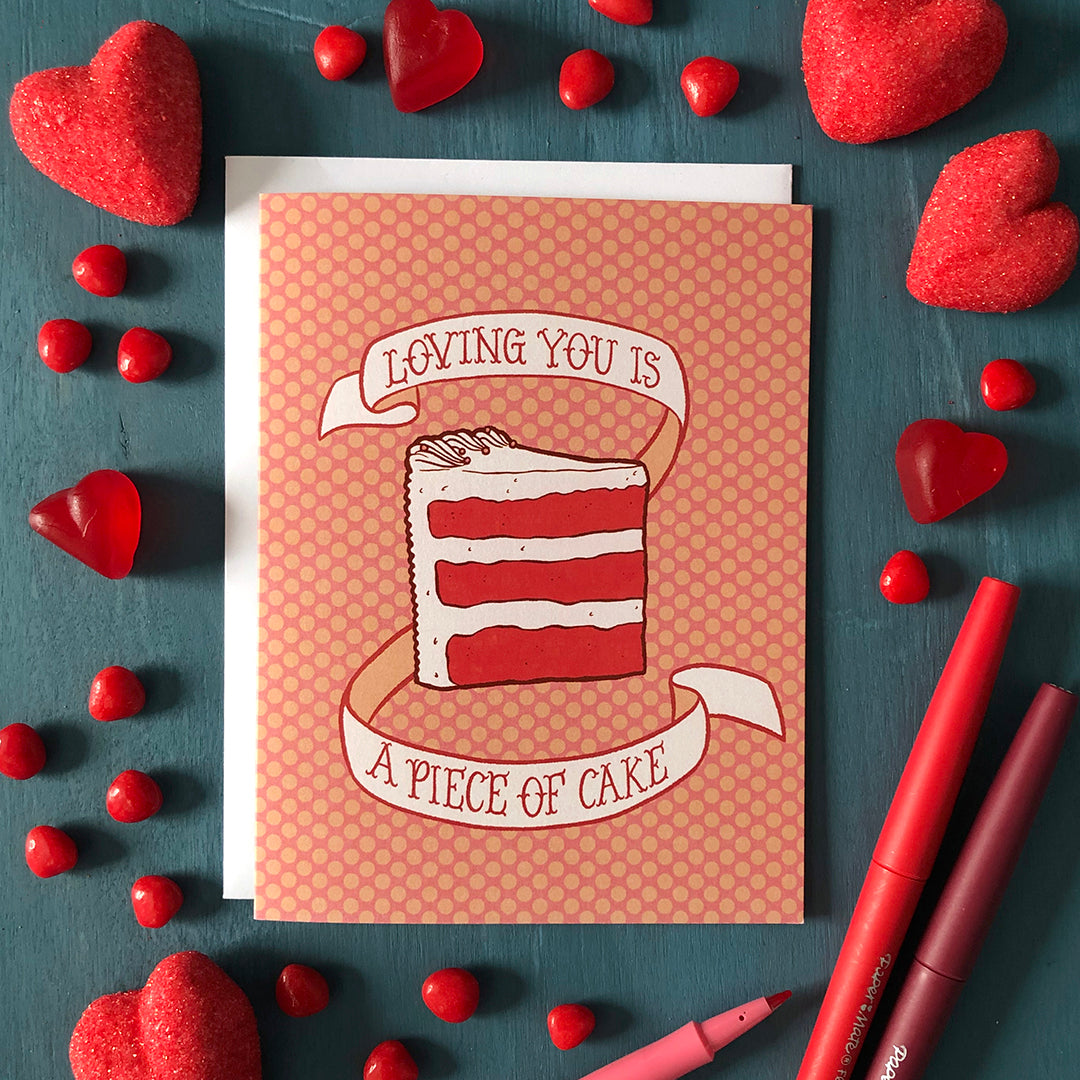 A pink polka dot greeting card shows an illustration of a triple-layer slice of red velvet cake and a banner that reads loving you is a piece of cake in hand lettering. The card is surrounded by pens and heart-shaped candy in various shades of red on a blue wooden background.