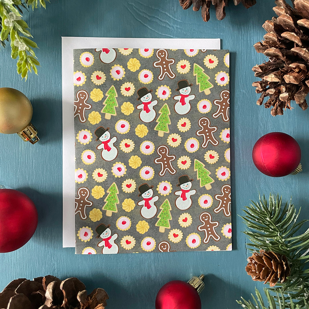 A greeting card is patterned with a variety of Christmas cookies - gingerbread men, snowmen, sugar cookie trees, empire cookies and shortbread. The card is on a blue background surrounded by ornaments, pinecones and faux greenery.