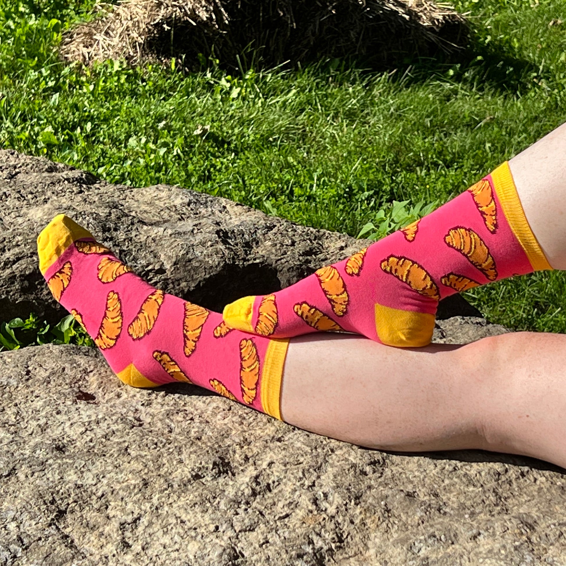 A pale, freckled pair of legs wears pink socks patterned with croissants with yellow cuffs, heel and toe. The legs are against a rock in a grassy background.
