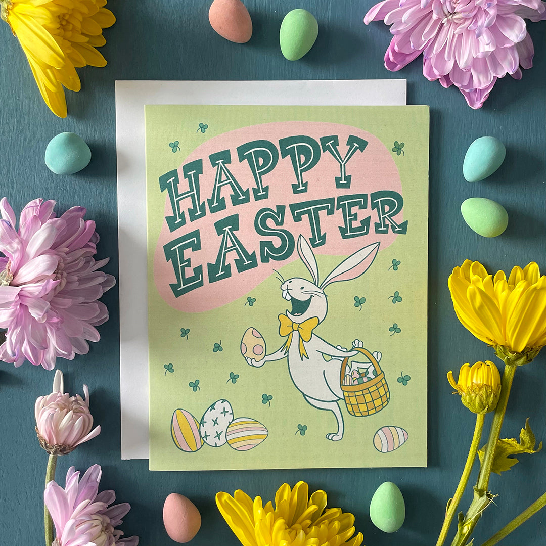 A pale green greeting card shows a cheerful white rabbit wearing a yellow bow tie holding a polka dotted easter egg in one paw and a basket of eggs in the other. There are easter eggs in front of and behind the rabbit. The card reads "Happy Easter" in midcentury-inspired hand-lettering and shows a shower of green clover. The card is surrounded by chocolate mini eggs and flowers.