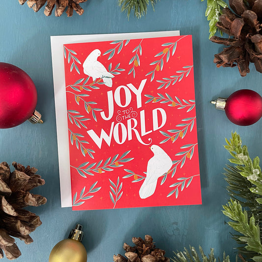 A red greeting card shows two white doves, branches of golden olives, and the hand-lettered words Joy to the World in white. The card is surrounded by ornaments, pinecones, and faux greenery.