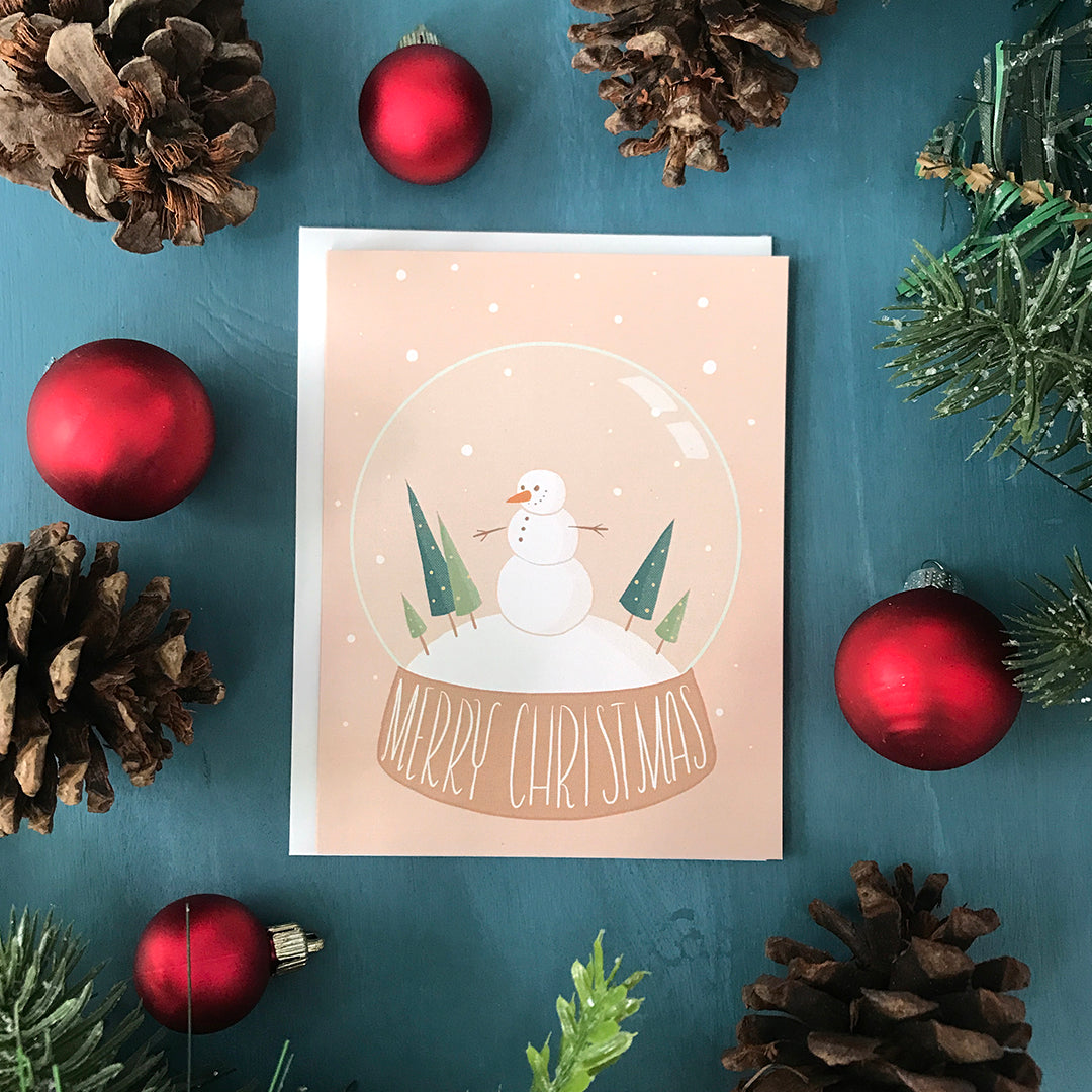 A pink Christmas card features a snow globe with a snowman, evergreens, and the words Merry Christmas lettered at its base. Snow dots fall both inside and outside the snow globe. The card is surrounded by pinecones, ornaments, and faux greenery.