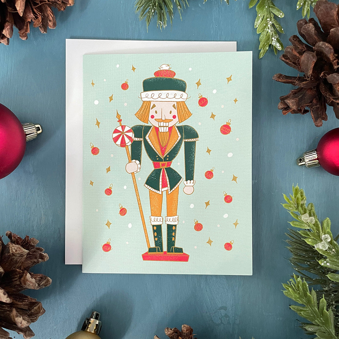 A mint card features a midcentury-style nutcracker in green and yellow, flanked by dots, yellow diamonds and red baubles. The card is surrounded by ornaments, pinecones and faux greenery.