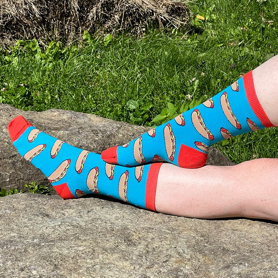 A pair of pale freckled bare legs wears blue socks patterned with hot dogs in buns and with red cuffs, heels and toes. The photo was taken outdoors against a grassy background.