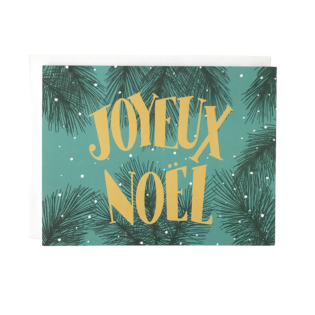 A teal greeting card features the words Joyeux Noel in midcentury-inspired yellow lettering against a background of snowy evergreen branches. The card sits against a white envelope on a white background.