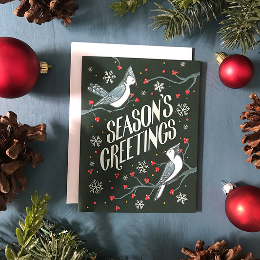 A dark green holiday card shows the words Season's Greetings. Around the letters are two blue jays perched on branches with red berries. The card is on a blue background surrounded by Christmas ornaments, faux greenery, and pinecones.