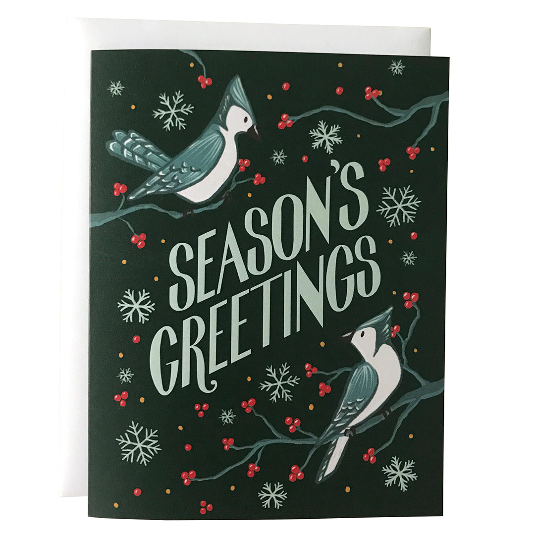 A dark green holiday card shows the words Season's Greetings in all capital letters. Around the letters are two blue jays perched on branches with red berries, along with some loose red berries and snowflakes. The card sits against a white envelope on a white background.