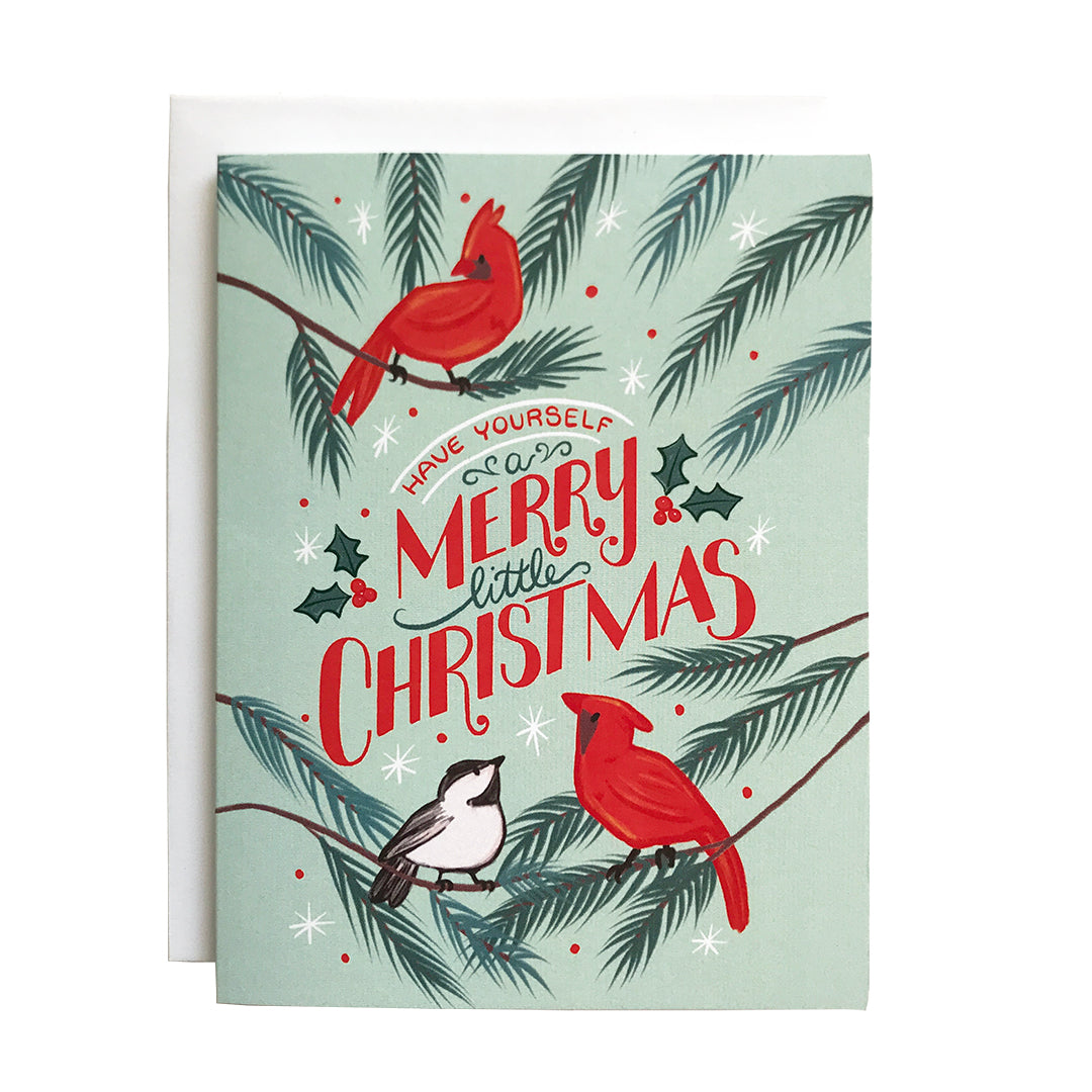 A greeting card with the words "Have Yourself a Merry Little Christmas" lettered in red and green, surrounded by red cardinals and a chickadee on a mint background. The card sits against a white envelope on a white background.