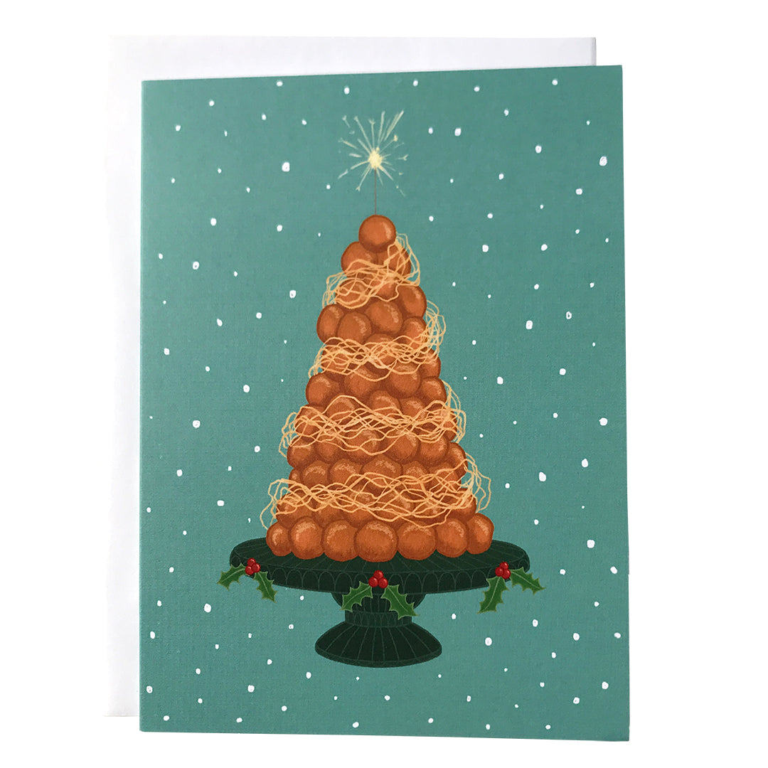 A light blue card shows a tall croquembouche sitting on a green, holly-laden pedestal platter. A sparkler is lit at the top of the dessert. The card is against a white envelope on a white background.