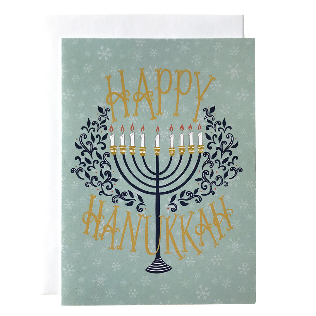 A pale blue snowflake-patterned greeting card reads Happy Hanukkah in deep yellow hand-lettering. In between the words is a navy menorah with lit candles flanked by navy flourishes. The card sits against a white envelope on a white background.