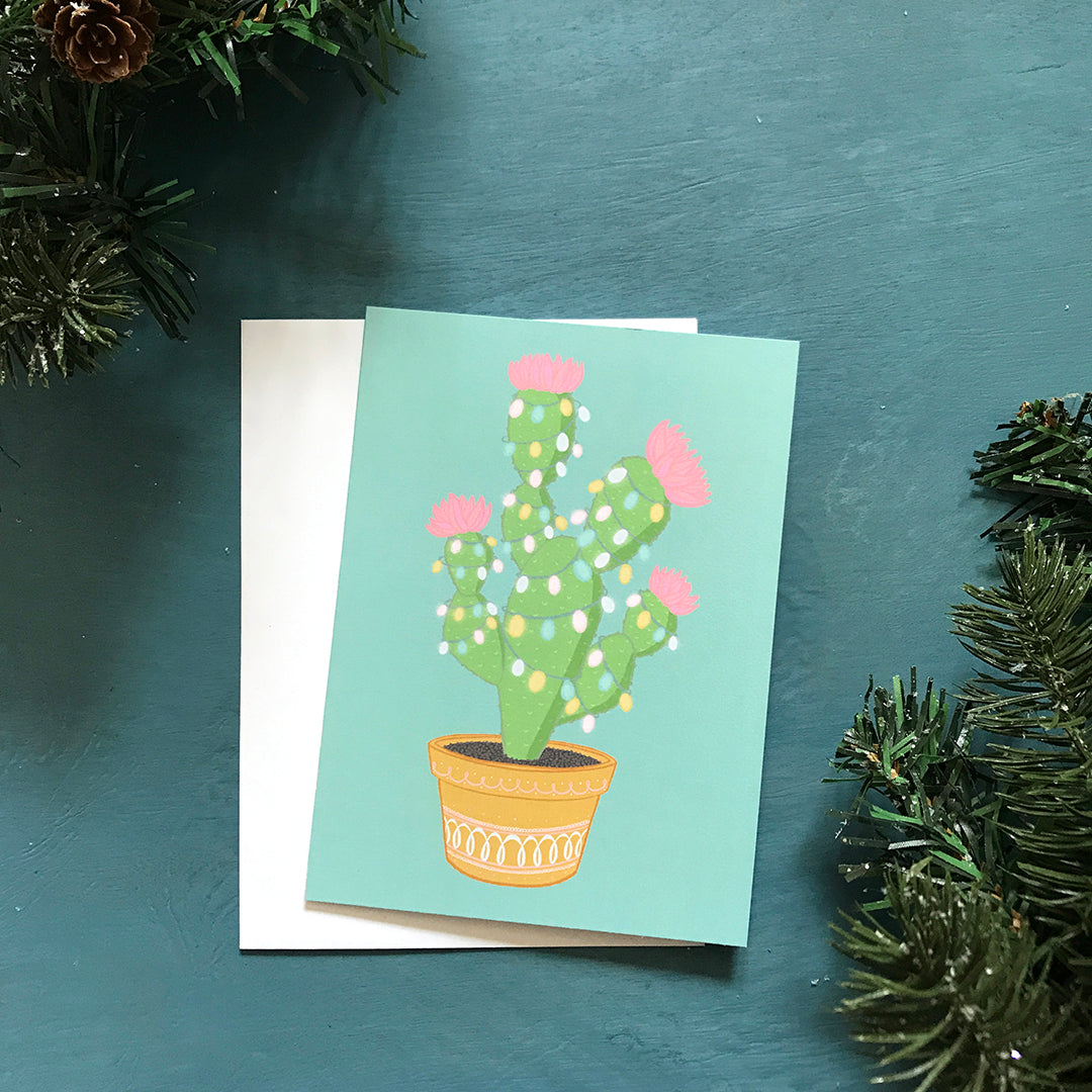 A light blue card features a potted green cactus with pink flowers strung with pastel lights. The card is surrounded by faux greenery.