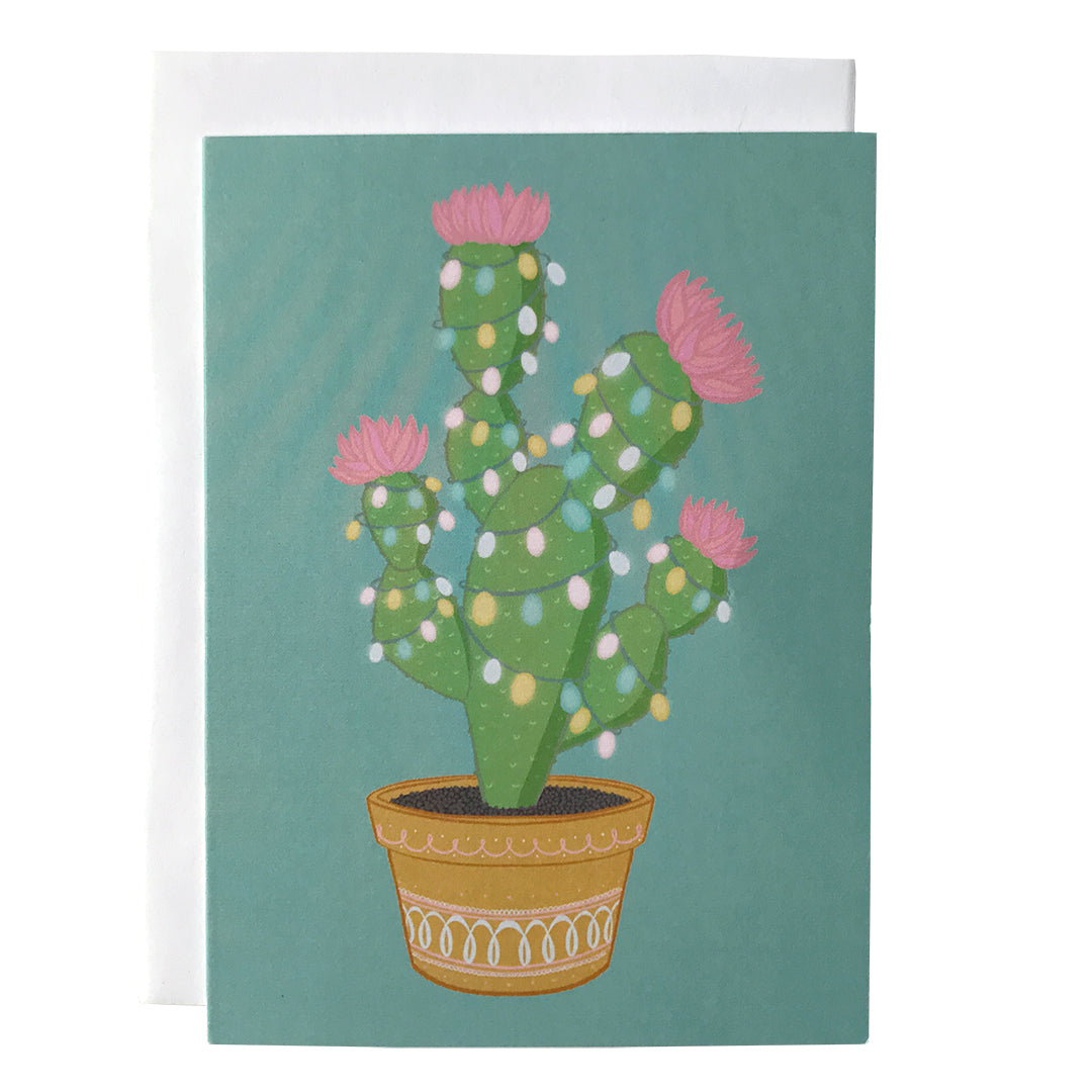 A light blue card features a potted green cactus with pink flowers strung with pastel lights. The card is on a white envelope against a white background.