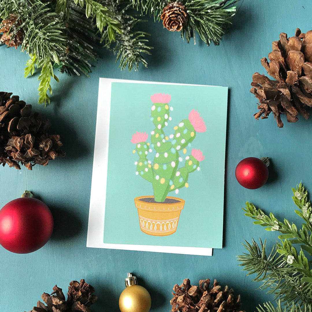 A light blue card features a potted green cactus with pink flowers strung with pastel lights. The card is surrounded by pinecones, ornaments and faux greenery.