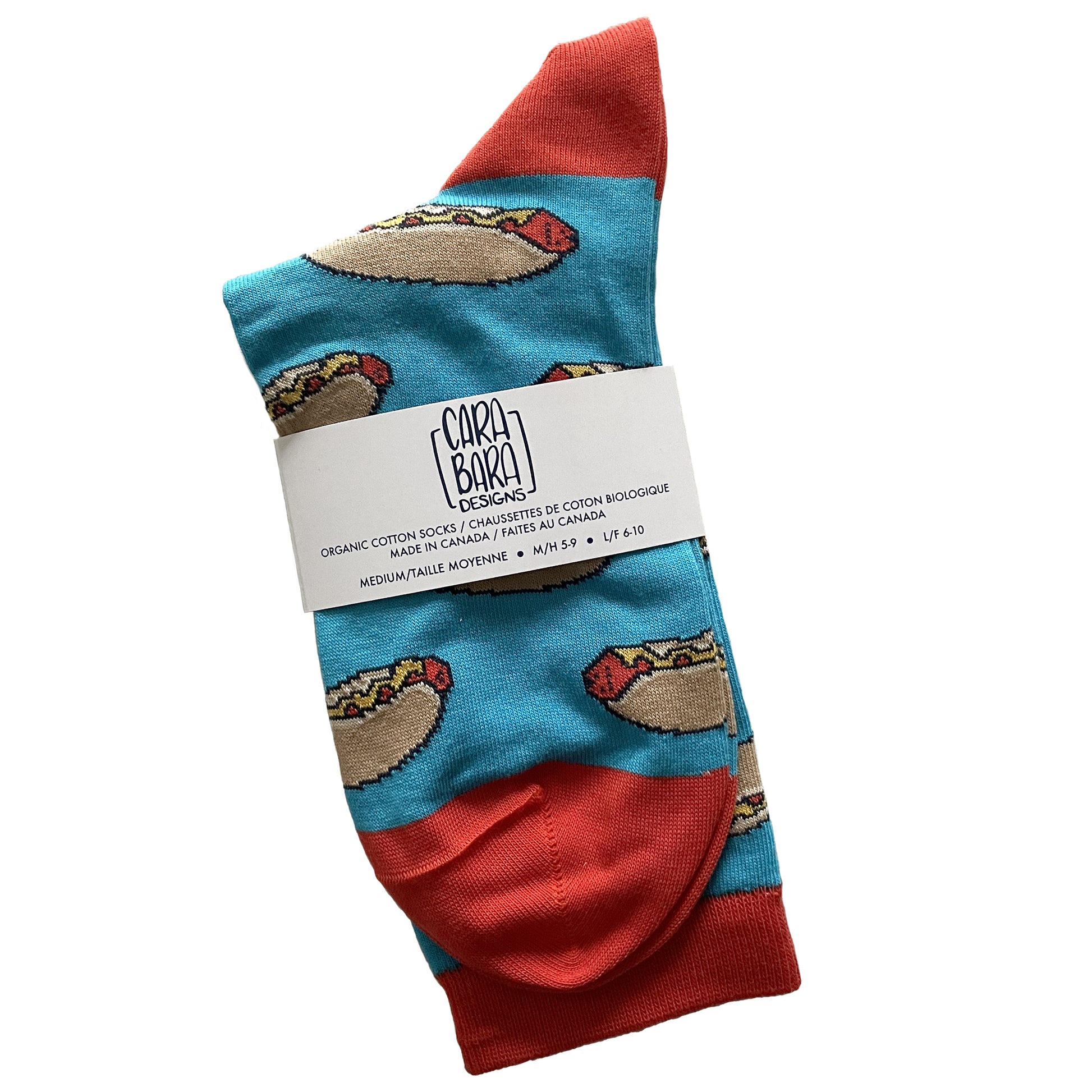 A pair of blue socks is patterned with hot dogs and has red accents. It is folded and labelled with a belly band with the Carabara Designs logo and the words organic cotton socks made in Canada, size medium, in English and French.