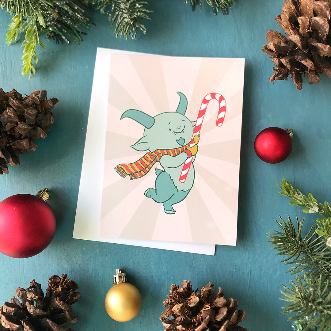 A pink card features a cute horned demon wearing mittens and a scarf and hugging a candy cane. The demon stands on two hooved legs. The card is flanked by pinecones, ornaments and faux greenery.