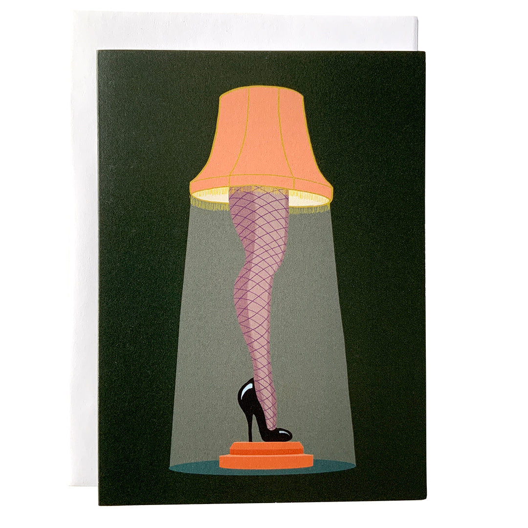 A dark green greeting card features an illustration of a lampshade atop a leg clad in fishnet stockings with a black high heel. The card sits against a white envelope on a white background.