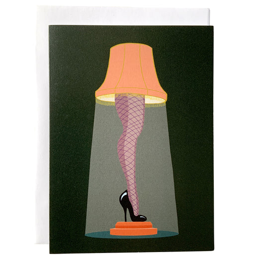 A dark green greeting card features an illustration of a lampshade atop a leg clad in fishnet stockings with a black high heel. The card sits against a white envelope on a white background.