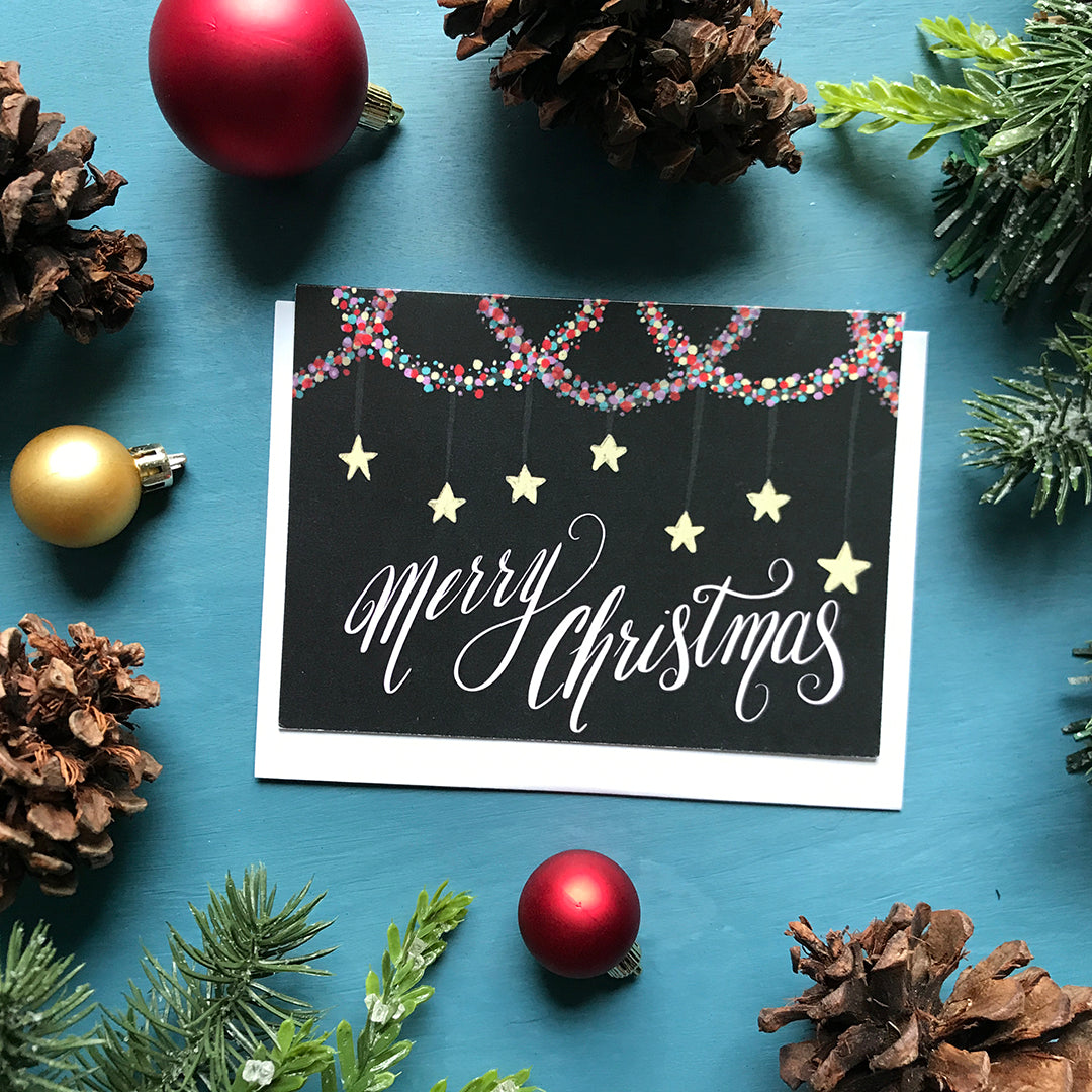 A black landscape-oriented greeting card shows the words Merry Christmas in pale pink with multi-coloured garland at the top and stars hanging above the words. The card is flanked by ornaments, pinecones and faux greenery.
