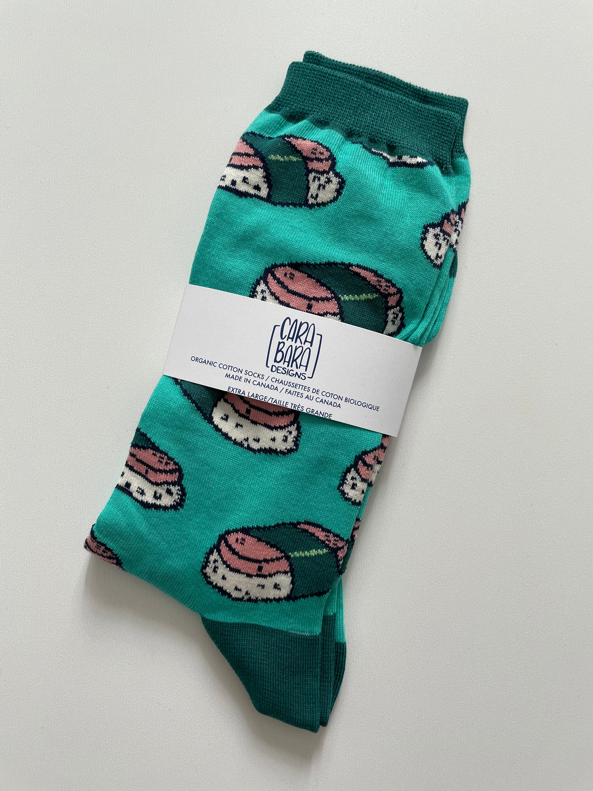A pair of turquoise socks with green cuffs and patterned with pink and green musubi is folded, packed labelled with the Carabara Designs logo, and the words organic cotton socks, made in Canada, extra large, in English and French.