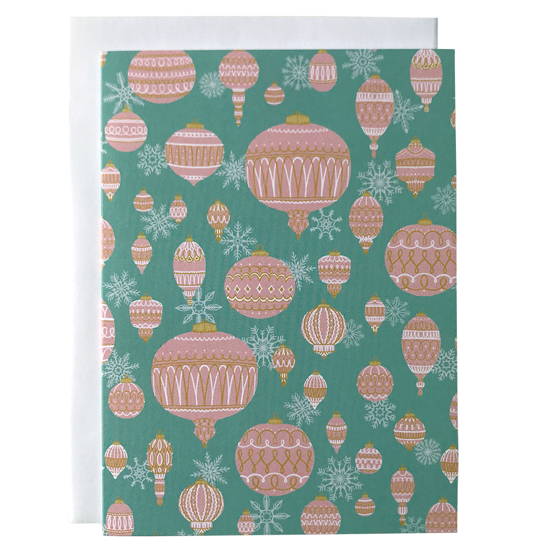 A greeting card is patterned with an assortment of pink ornaments and white snowflakes on a light aqua blue background. The card is against a white envelope on a white background.