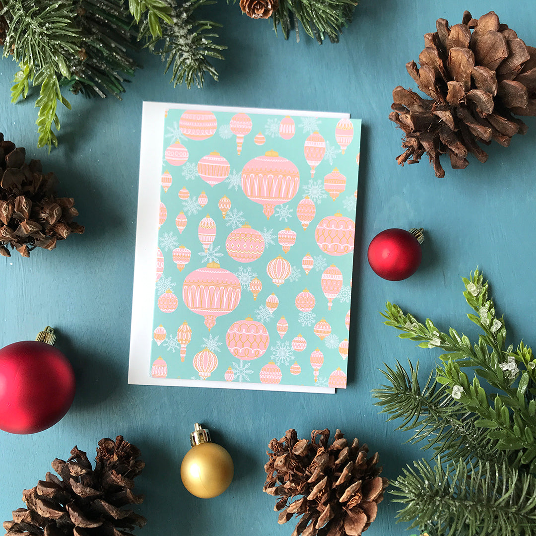 A greeting card is patterned with an assortment of pink ornaments and white snowflakes on a light aqua blue background. The card is flanked by ornaments, pinecones and faux greenery.