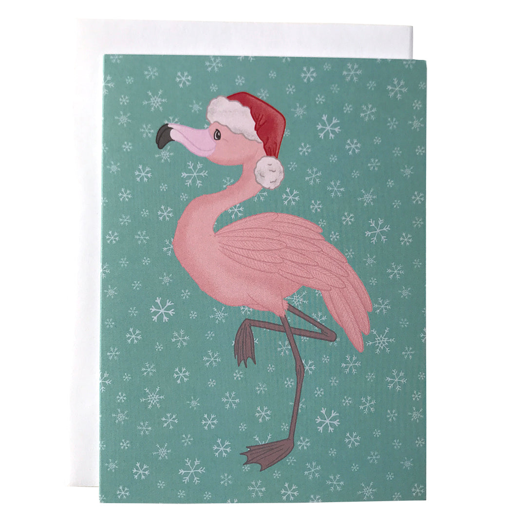 A light blue snowflake-patterned card features a pink flamingo wearing a Santa hat and standing on one leg. The card is against a white envelope on a white background.
