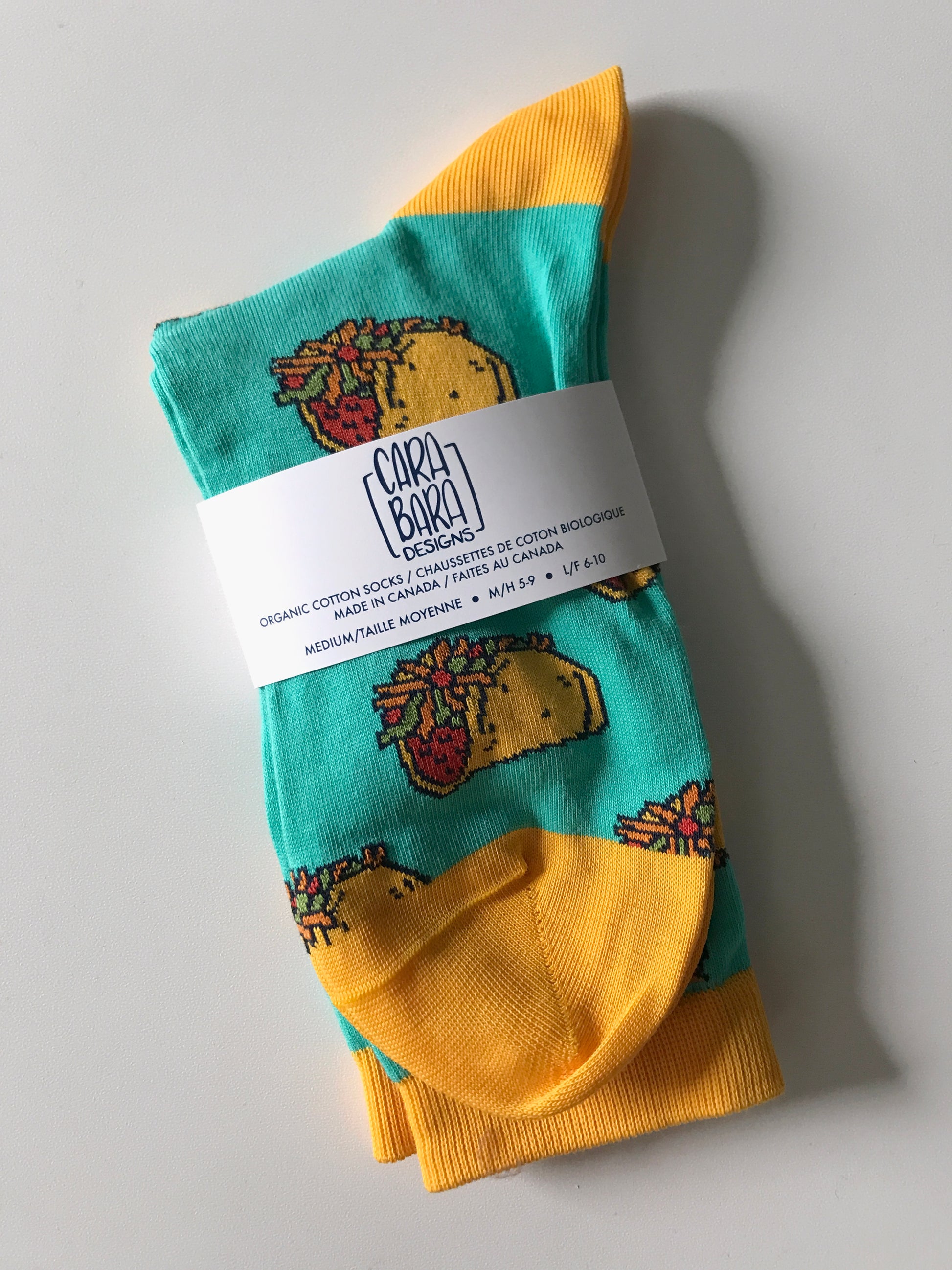 A pair of aqua-blue socks with yellow accents is patterned with tacos and is folded and labelled with a belly band with the Carabara Designs logo and the words organic cotton socks made in Canada, size medium, in English and French.
