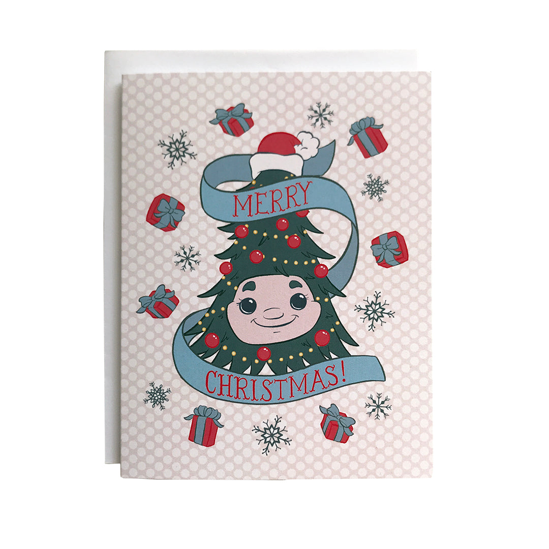 A Christmas card features an illustration of Woody the Christmas Tree, some gifts with bows and blue snowflakes on a pink dotted background. A banner reads Merry Christmas in hand-lettering. The card sits on a white envelope on a white background.