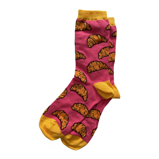 A pair of pink socks is patterned with orange-and-yellow croissants with dark brown outlines. The cuffs, heels, and toes of the socks are yellow.